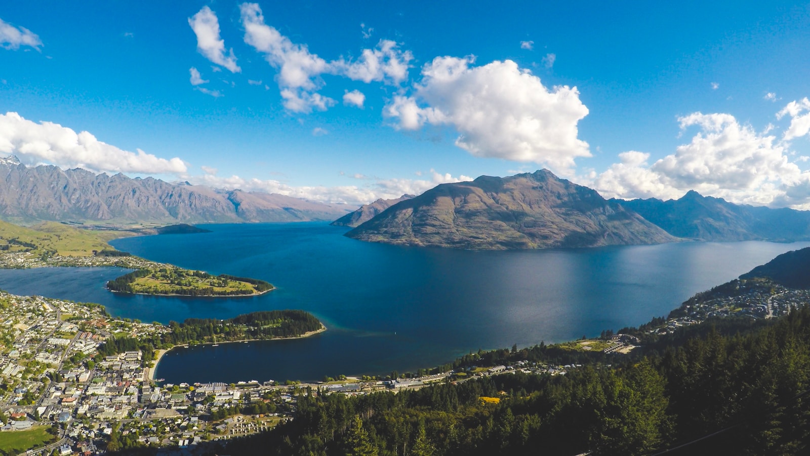 Des bâtiments près d'un plan d'eau et des montagnes sous un ciel bleu clair avec quelques nuages blancs en plein jour.